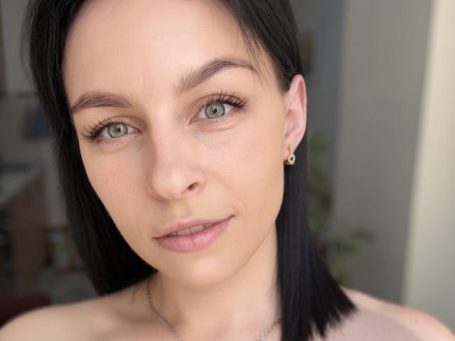 EmmaNoa hat eine helle Haut, grüne Augen, schwarzes, glattes Haar, kleine Ohrringe und ein zartes Lächeln. Sie ist oben ohne, hat eine kleine Nase und volle, rosafarbene Lippen..