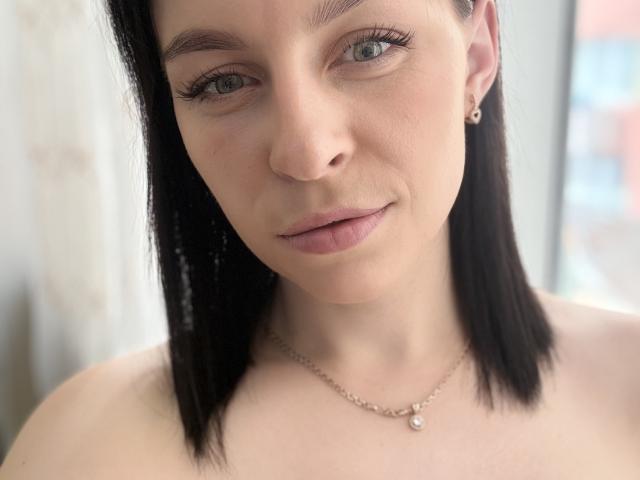 EmmaNoa mit schwarzen, glatten Haaren, blauen Augen, heller Haut, dezentem Make-up, rosafarbenen Lippen, kleinen Ohrringen, einer Halskette mit einem kleinen Anhänger und bloßen Schultern in einem Selfie bei weichem Licht..