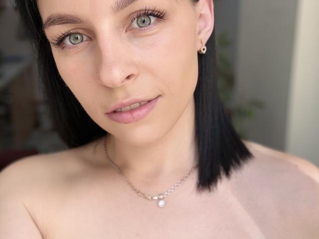 EmmaNoa met bleke huid, groene ogen, zwart steil haar, kleine oorbellen, een subtiele ketting, geen top aan, lichtroze lippen die iets open zijn, een zachte glimlach en subtiele make-up, op een selfie..