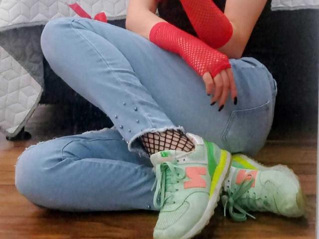 Stephanie Sun sentada com as pernas cruzadas, vestindo jeans azuis, luvas de meia-calça vermelhas, unhas pretas, tênis brancos com detalhes rosa e verde, e meias de meia-calça rasgadas, em um piso de madeira..
