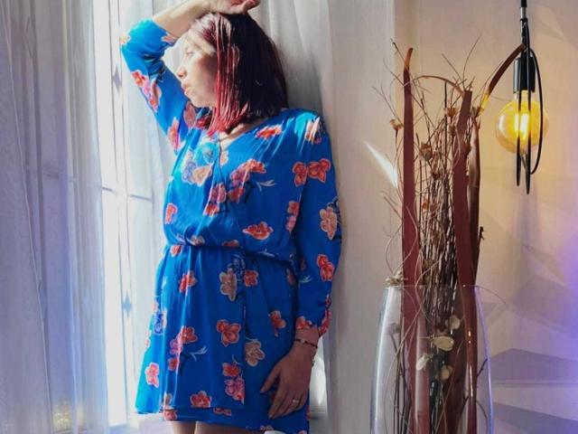 Stephanie Sun, in un abito blu con fiori, in piedi vicino a una finestra, con un braccio alzato, la luce del sole sui capelli rosso-marrone, che guarda fuori, accanto a un vaso alto con canne secche e una lampada..
