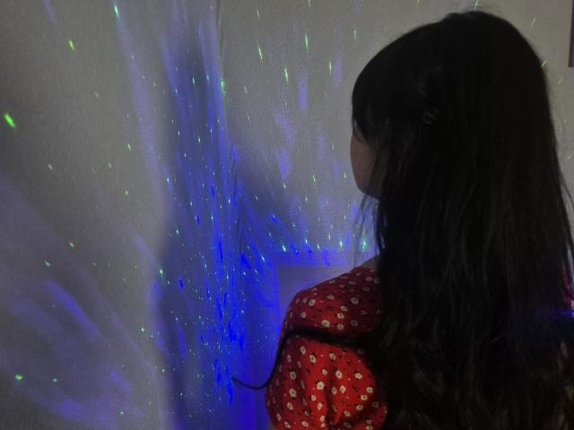 WenTina with long black hair, wearing a red polka-dot shirt, stands with her back to the camera, looking at colorful green and blue light patterns on a wall.
