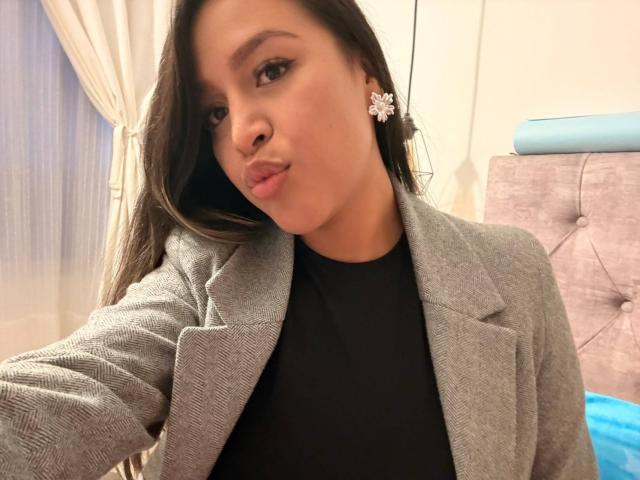LiaDiane, com cabelos castanhos longos, vestindo um blazer cinza sobre uma blusa preta, com lábios curvados para baixo e brincos florais, olhando para a câmera. Seus seios são pequenos..