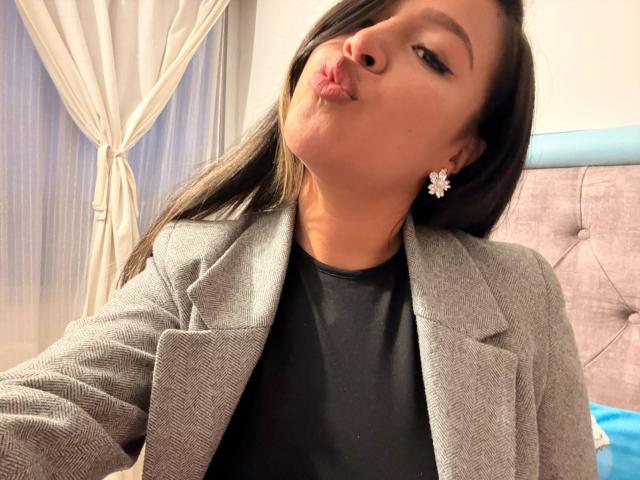 LiaDiane, com os lábios curvados, usando um blazer cinza, uma camisa preta e brincos de flores, olhando para o lado, com cabelo castanho escuro, pele clara, com uma expressão adorável, em um quarto aconchegante..