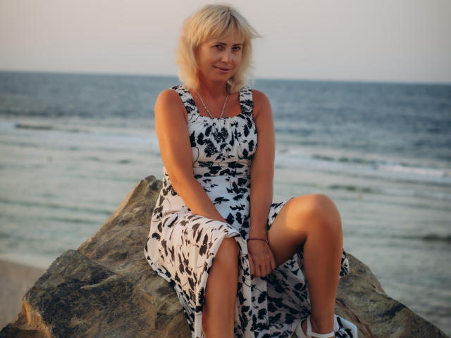 Lauren Wishy, met blond haar, draagt een zwart-wit bloemenjurk, zit op een rots, met haar knieën samengevouwen, en glimlacht naar de camera. Op de achtergrond is de oceaan en golven te zien..
