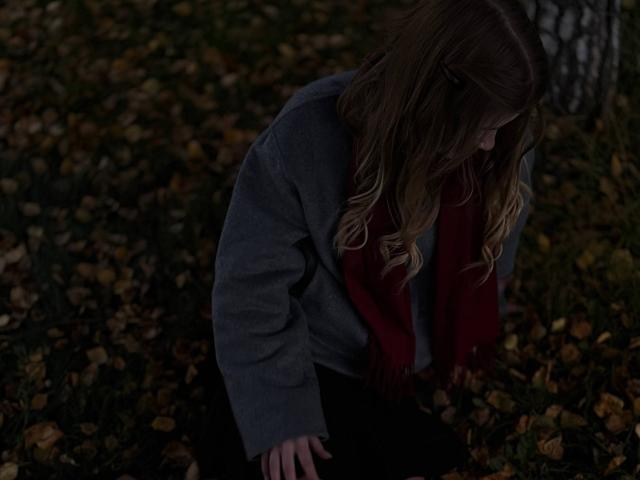 Norah Habrock, con su largo cabello castaño ondulado, un suéter gris, una bufanda roja y pantalones negros, está agachada en el suelo oscuro cubierto de hojas, mirando hacia abajo con la cabeza ligeramente inclinada..