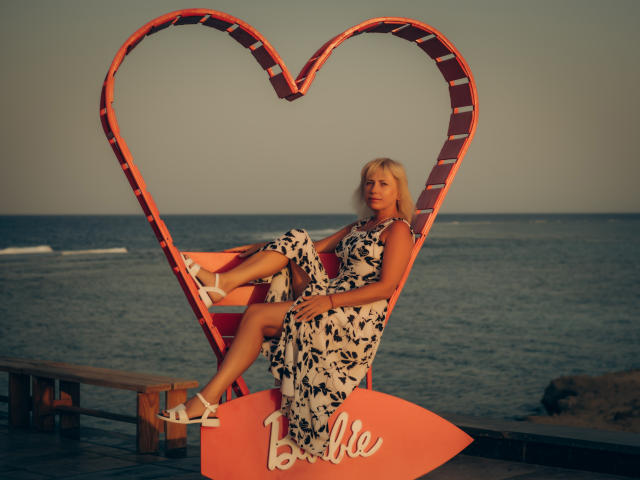 Lauren Wishy, com cabelo loiro, vestindo um vestido floral branco e preto, sentada em uma estrutura laranja em formato de coração, exibindo suas nádegas, com saltos brancos, à beira-mar ao pôr do sol..