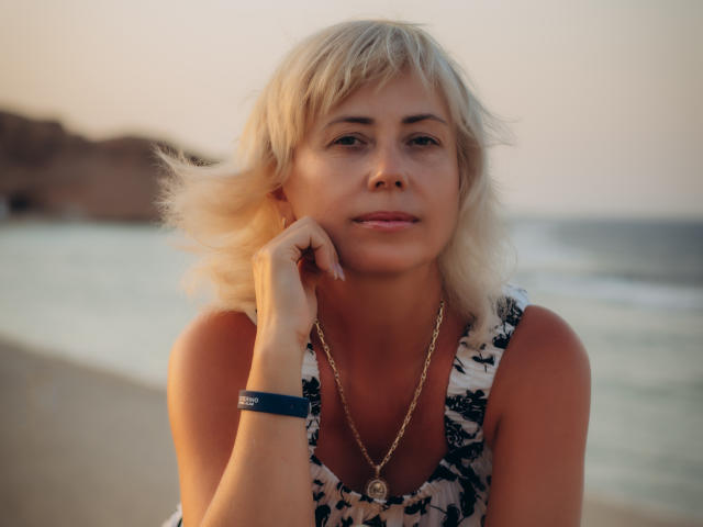 Lauren Wishy, con i capelli biondi, indossa un abito floreale, una collana dorata e un braccialetto blu, appoggia il mento sulla mano e guarda leggermente di lato, in spiaggia durante il tramonto..
