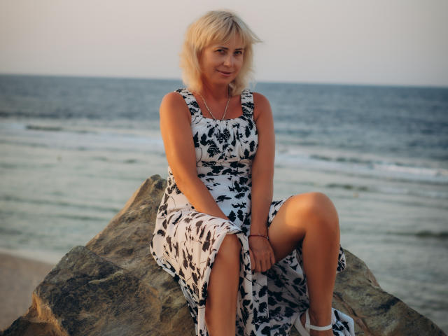 LaurenWishy, blond, witte jurk met zwarte patronen, zittend op een rots, benen samengevoegd, lichte glimlach, oceaan op de achtergrond, zonsondergang, handen op de knieën, casual, schattig, subtiele make-up, ontspannen..
