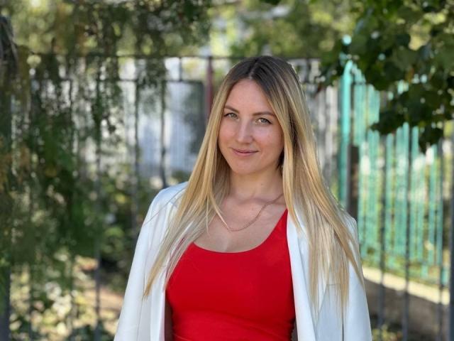 JuicyTigerss a les cheveux blonds, porte un haut rouge et une veste blanche, et se tient dehors avec un sourire discret, portant un collier fin et ayant une allure mignonne..