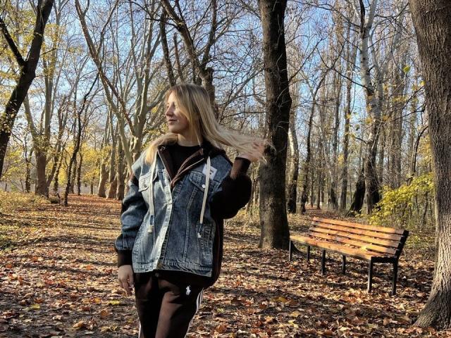JuicyTigerss with long blonde hair, wearing a denim jacket over a black top and brown pants, stands in a forest, touching her hair, surrounded by fallen leaves and trees.