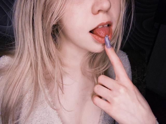 TiffaniMacandog's face, tongue out, licking a purple nail with her right index finger, blonde hair, pale skin, and a soft beige sweater, showing a sexy, flirty expression.