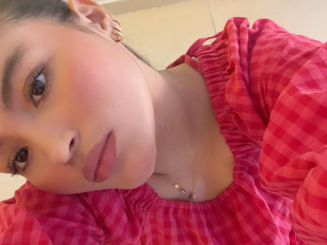 MiiaPalmer with pink checkered shirt, puffy sleeves, brown eyes, full lips, pink lipstick, necklace, ear piercings, light skin, dark hair in a bun, looking at camera.