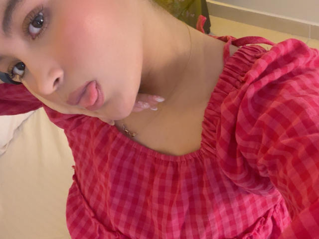 MiiaPalmer with pink checkered dress, tongue out, puffy sleeves, light brown skin, pink lipstick, and a necklace, in a selfie. Boobs slightly visible under dress.