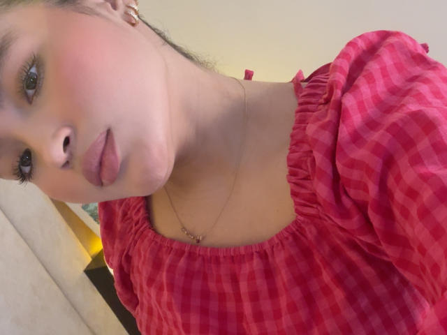 MiiaPalmer with pouty lips, wearing a pink checkered top with ruffled shoulders, and a thin necklace, has fair skin and brown eyes, looking at the camera.