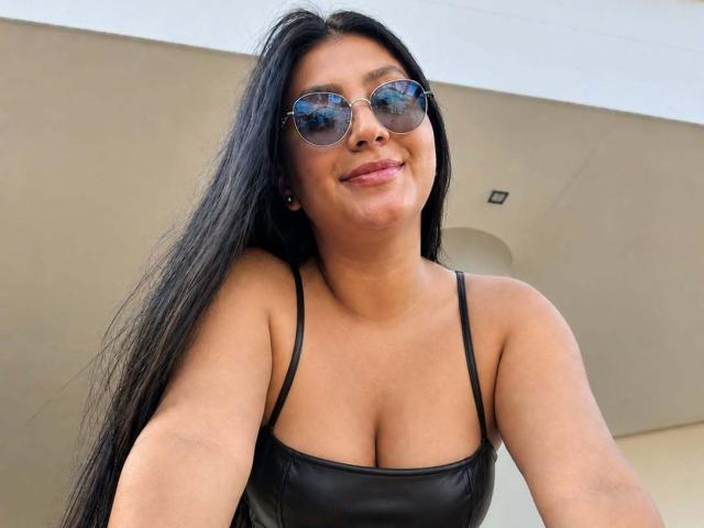 Catalina Rivero, con lunghi capelli neri, indossa occhiali da sole rotondi e una giacca di pelle nera, che mette in mostra il seno e sorride leggermente. Ha la pelle abbronzata e piccoli orecchini..