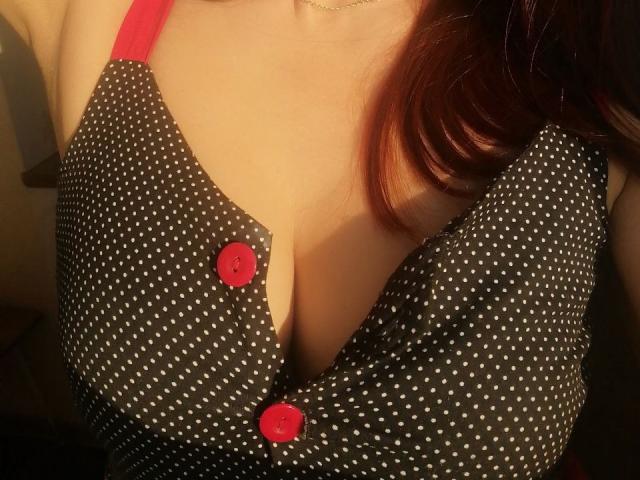 AlexisLovelyy's polka-dotted dress with red buttons, showing ample cleavage, has red straps and is slightly open, highlighting her large, rounded tits. Her red hair is visible.