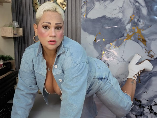 GianellaCox met een platinablonde pixie-kapsel, een outfit van blauwe denim, witte hoge hakken, grote borsten en roze blush, op handen en knieën, kijkend naar de camera, in een moderne kamer..