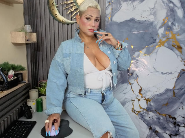 GianellaCox met kort blond haar, die een blauwe denimjas draagt, een wit topje met strikjes dat haar grote borsten laat zien, en versleten jeans, terwijl ze een telefoon vasthoudt en aan een bureau zit..
