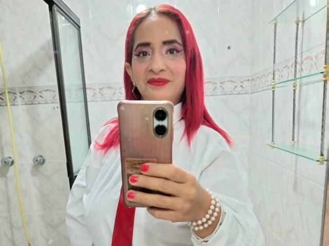 KateMonroe with pink hair, white shirt, red tie, and red lipstick, taking a mirror selfie with a rose gold phone, wearing a pearl bracelet, in a bathroom.