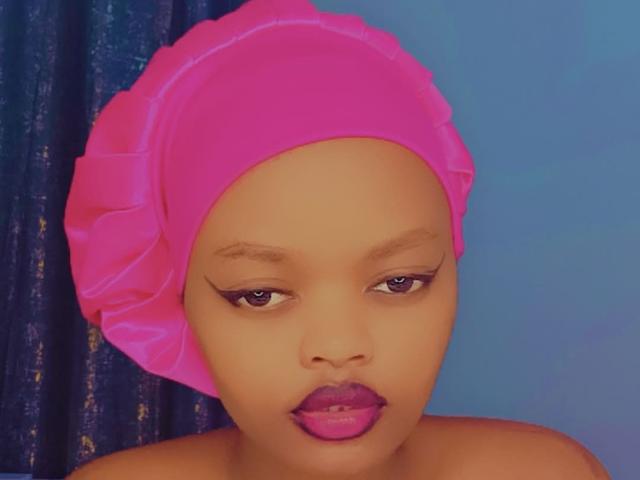 NaughtyPatricia with a pink headscarf, purple eyeliner, and purple lipstick, looking down with a serious expression, has smooth brown skin and a cute face.