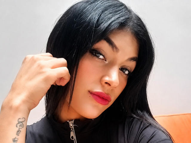 AlexandraBuby has black hair, brown eyes, red lipstick, and a lip piercing. She's wearing a black top with a zipper, touching her hair, showing a tattoo on her wrist.
