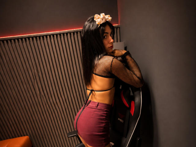  AlexandraBuby with long black hair, wearing a mesh top, burgundy shorts, and a flower hairpiece, leaning on a chair, showing her ass and back, looking over her shoulder.