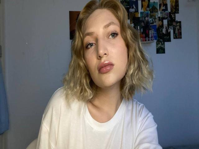 SigneScafuri, met golvend blond haar, draagt een witte T-shirt, heeft roze lippen die ze opvouwt en kijkt recht in de camera. Haar achterkant is niet zichtbaar. Haar borsten zijn klein..