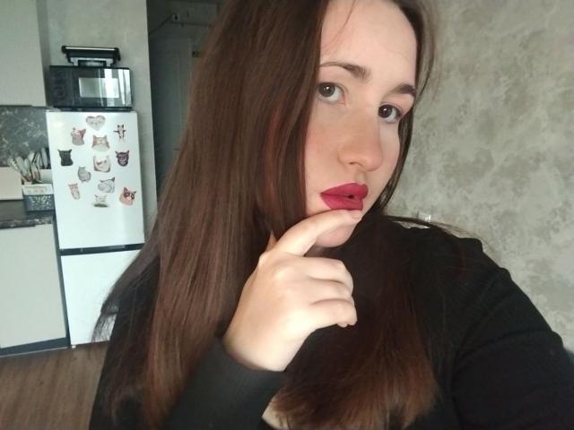 RosalindCutsinger with long brown hair, red lipstick, and black top, sucking on her finger, looking at the camera, in a kitchen with a white fridge and microwave.