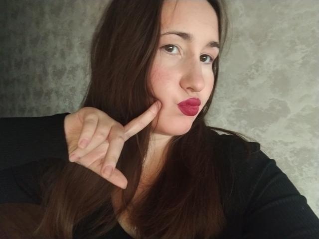 RosalindCutsinger has long brown hair, dark lipstick, and is pouting with her finger on her cheek, wearing a black top, against a textured gray wall.