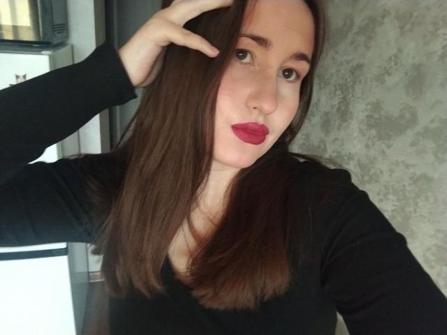 RosalindCutsinger, com cabelos castanhos longos, blusa preta e batom vermelho, fazendo uma expressão de lábios franzidos, com a mão tocando a cabeça, e uma parede cinza texturizada ao fundo..