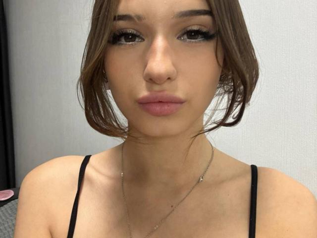ShanekaKonig hat ein hübsches Gesicht mit braunen Augen, vollen, rosafarbenen Lippen und braunen Haaren, die zu einer unordentlichen Seitenpartie gestylt sind. Sie trägt ein schwarzes Top mit Trägern und eine dünne Halskette..