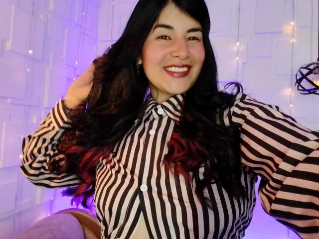 NinaHarpper with long black hair, striped shirt, and red ombre, smiling, hand in hair, showing her tits, cute and sexy, purple light in background.