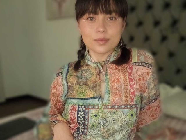 Iniridax with puffy brown eyes, bangs, and braided hair, wearing a colorful, patterned, quilted shirt, showing her tits slightly under the fabric.