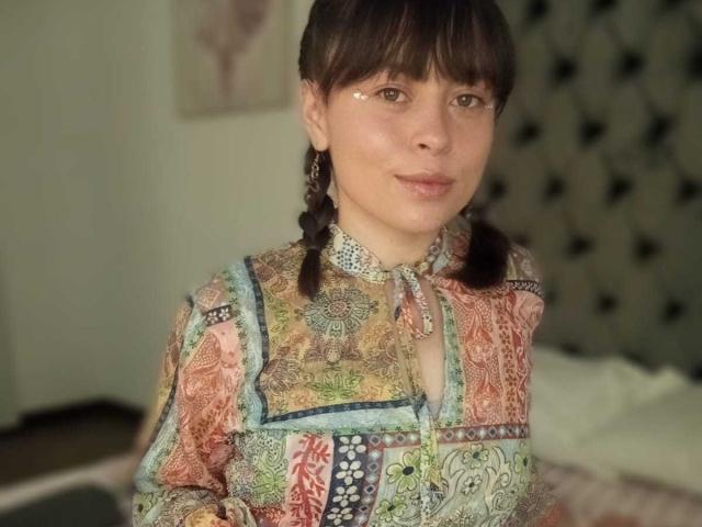 Iniridax with brown pigtails, wearing a colorful, intricate patterned dress, has a gentle smile, and a slight tear on her right cheek, standing in a softly lit room.