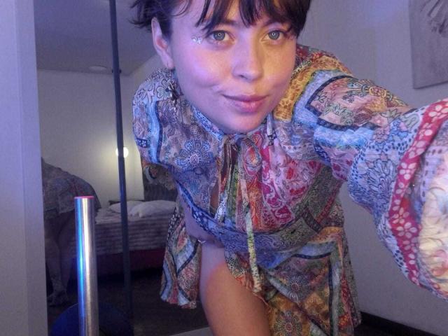 Iniridax with short brown hair and blue eyes, wearing a colorful, patterned dress, pouting with a slight smile, showing a thigh and no pants, in a bedroom.