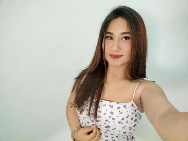 Julhiana mit langen, braunen Haaren, trägt ein weißes, blumiges Crop-Top, das ihre kleinen Brüste zeigt, sie hat helle Haut und ein leichtes Lächeln und macht ein Selfie mit ihrer rechten Hand..