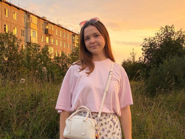 JeseniaPiontek with brown hair, pink tee, white floral skirt, white crossbody, pink sunglasses on head, standing in grassy field at sunset, orange sky, brick building in background.