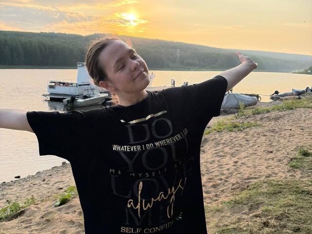 JeseniaPiontek stands on a sandy beach, arms out, wearing a black "Do Whatever I Want" t-shirt, smiling at sunset with a small boat in the background.