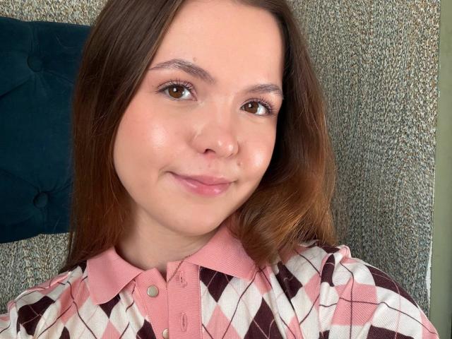 JeseniaPiontek with brown eyes, pink shirt, and diamond pattern, smiling softly. She has light skin, straight brown hair, and a cute, slightly flushed face. Her tits are small.