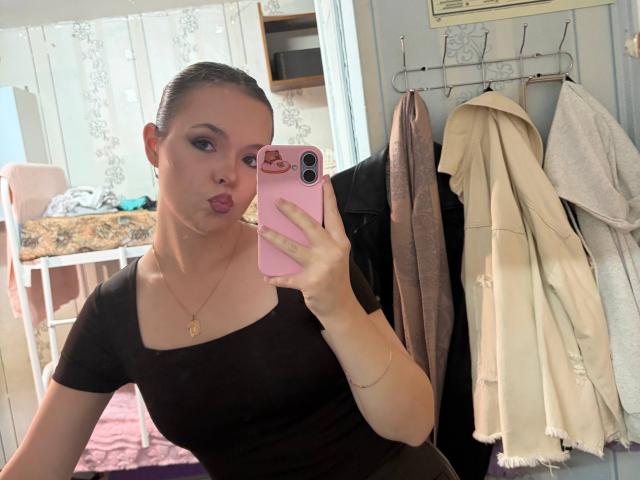 JeseniaPiontek with puffed lips, taking a mirror selfie in a bedroom. She's wearing a black shirt, gold necklace, and holding a pink phone. Her ass is not visible.