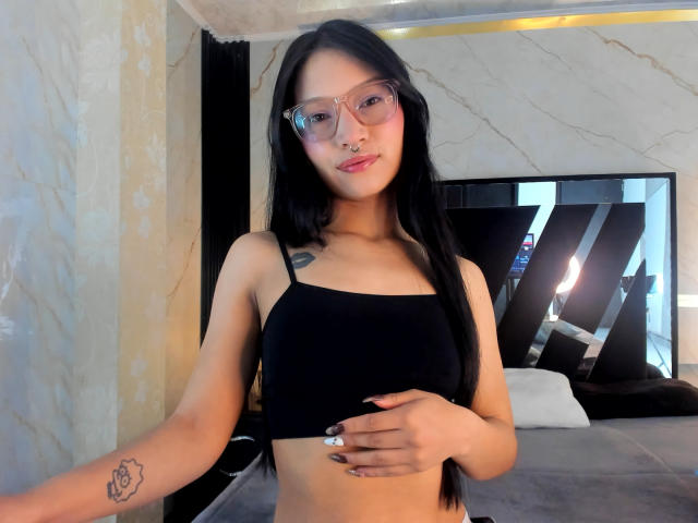 Sophie Delaney, con cabello negro largo, gafas y un top corto negro, sosteniendo sus pechos, mostrando un pequeño tatuaje en su brazo, y sonriendo en un dormitorio..