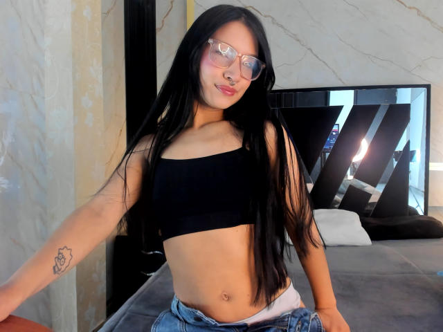 Sophie Delaney, avec ses longs cheveux noirs, portant des lunettes et un débardeur de sport noir, laissant apparaître son ventre plat et un petit tatouage sur son bras droit, est assise sur un lit..