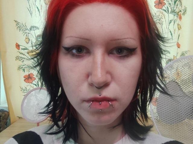 ParticiaFeiler, con capelli rosso-neri, piercing al naso, al labbro inferiore e al mento, indossa una camicia a righe nere e bianche, ha l'eyeliner nero e la pelle pallida..