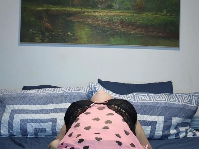 AngelaMonticellii è sdraiata su un letto, a faccia in giù, indossa un abito rosa con motivi a cuore e maniche in pizzo nero, che mostrano il suo sedere e la schiena..