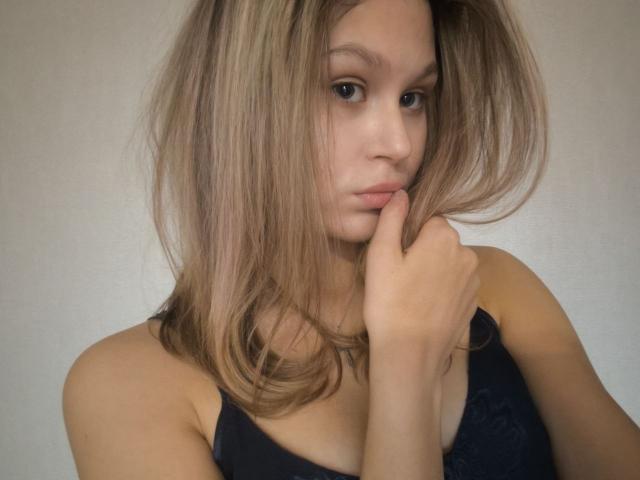 YungQuiller with blonde hair, pouty lips, and a finger on her chin, wearing a black tank top, looking slightly annoyed. Her tits are small, and she's cute.