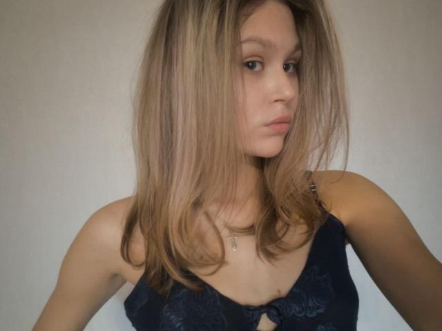 YungQuiller with blonde hair, wearing a dark blue lace top, has a serious expression, slightly parted lips, and a small cross necklace. Her tits are visible through the lace.