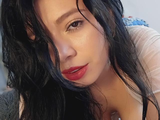 Linda Mancini, con i capelli neri bagnati, il rossetto rosso e gli occhi marroni, è piegata in avanti, mostrando il décolleté con una maglia trasparente. La sua pelle è liscia e ha un aspetto seducente..