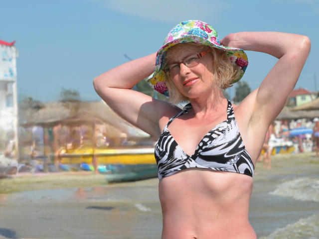SunnySoul-hot with short blonde hair, wearing a floral sunhat, zebra-print bikini top, and glasses, standing on a beach with arms raised, showing small tits and a tan line.
