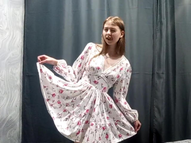 MargretWarriner in a white floral dress with pink roses, holding out the skirt, standing in a room, light brown hair, slight smile, background is gray curtains.